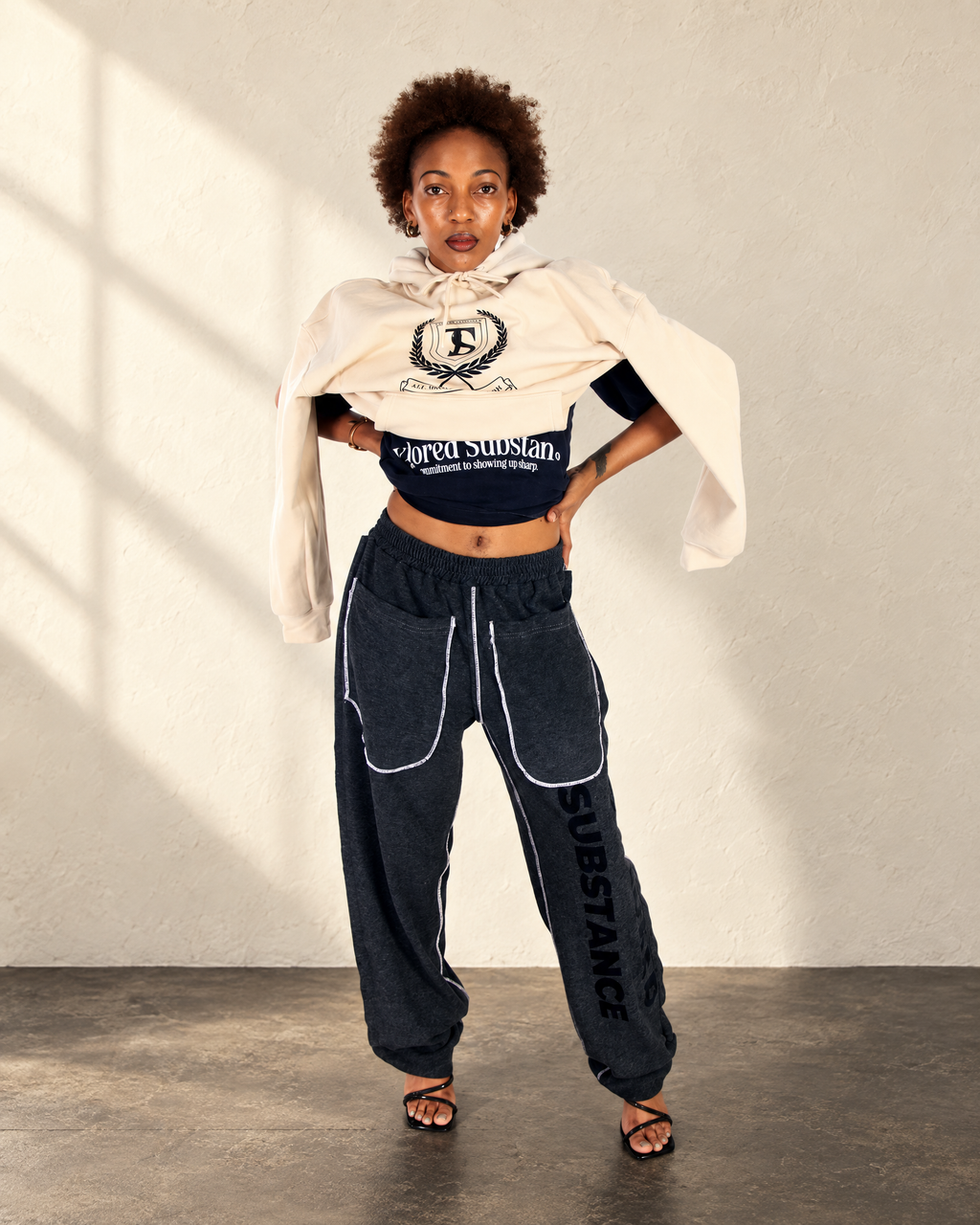 Model wearing the cream Crest Hoodie with a black embroidered crest over a dark top and charcoal trousers.