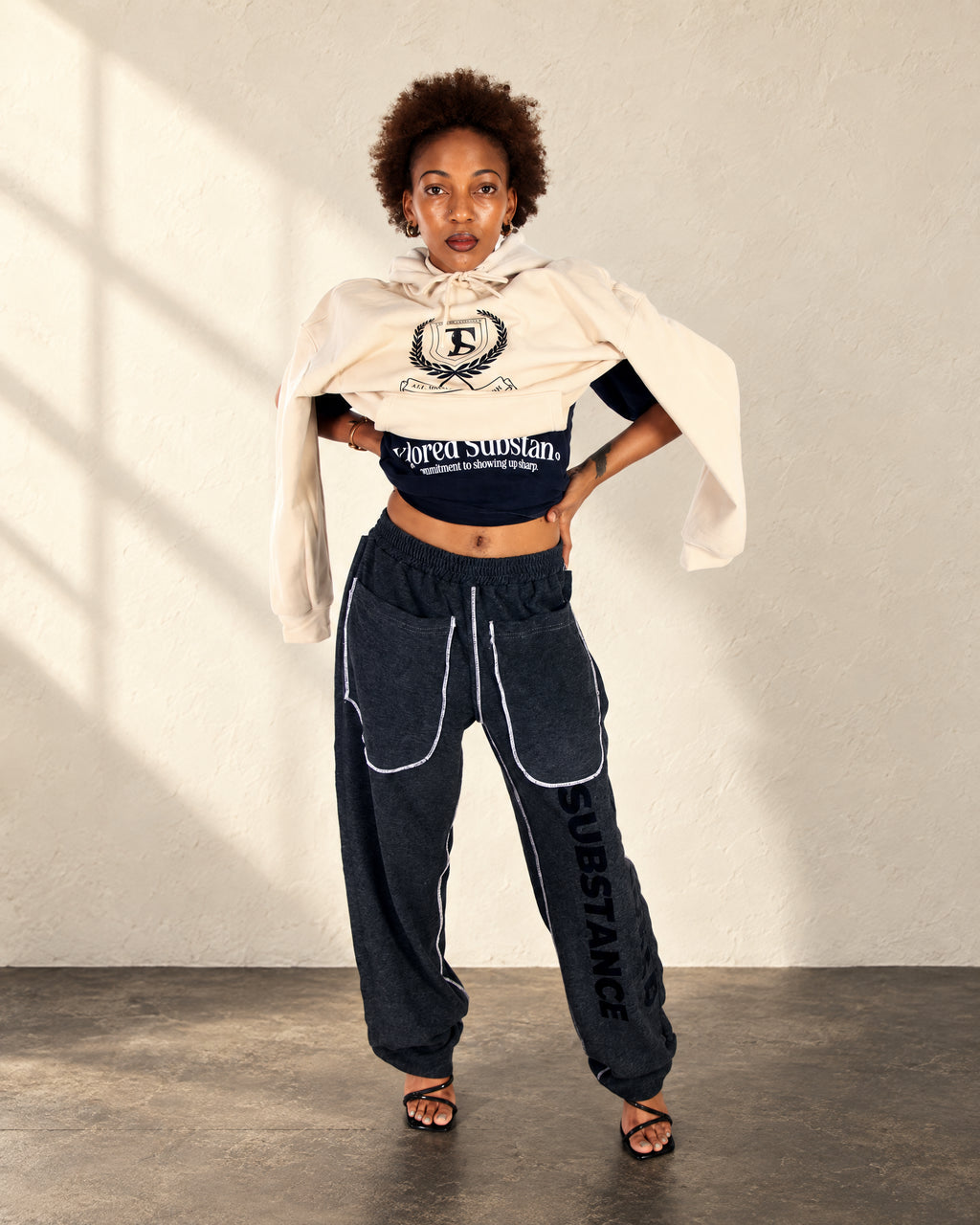 Model wearing the cream Crest Hoodie with a black embroidered crest over a dark top and charcoal trousers.